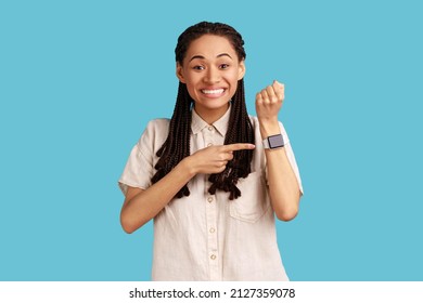 Portrait Of Happy Excited Woman With Black Dreadlocks Standing, Looking At Camera, Pointing At Her Smart Watch, Wearing White Shirt. Indoor Studio Shot Isolated On Blue Background.