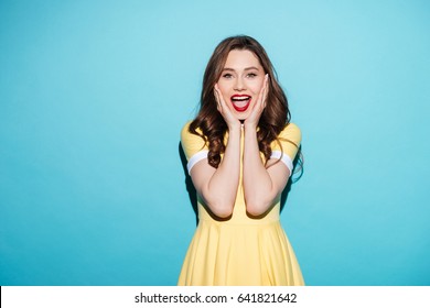Portrait of a happy excited woman in dress with open mouth keeping hands at her face isolated over blue background