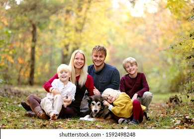 A portrait of a happy family of 5 people, inclluding three cute smiling children, sitting outside in the woods with their pet dog on an Autumn day.