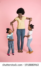 Portrait of happy family in colorful stylish outfits, charming young African woman mom and two cute little kids girls, having fun, playing and dancing together, on isolated pink background
