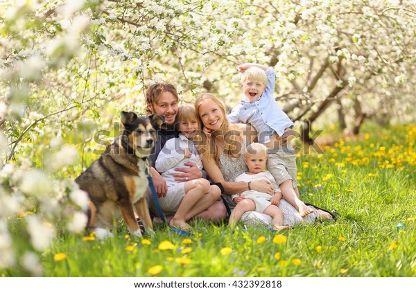 A portrait of a happy family of five people,\
including mother, father, 2 young children and baby girl and their\
pet dog hugging outside in a flower meadow under blossoming apple\
trees on a spring day.