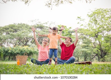 Portrait Of Happy Family Man Woman And Little Girl Playing Outside. Young Family Of Three Having Fun Picnic Together Outdoor. Asian Parents Girl Jumping Happy And Smile. Happiness And Harmony In Life.