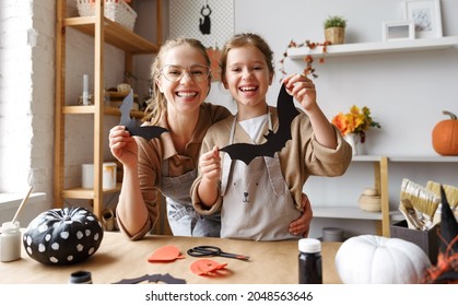 Portrait Of Happy Family Mother And Cute Girl Daughter With Cutted Black Paper Bats In Hands Smiling At Camera While Sitting At Wooden Table And Making Different Halloween Decor Together At Home