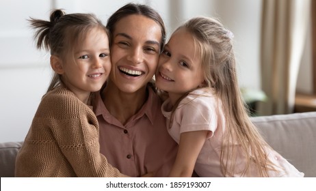 Portrait Of Happy Family, Mother Day Celebration Concept. Single Loving Mommy And Two Little 6s Daughters Embracing Seated On Sofa At Home. Young Cheerful Mom With Cute Girls Smile Looking At Camera