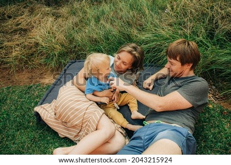 Portrait of happy family resting on grass outdoors. Couple lying on mattress and teasing their toddler daughter. Family vacation concept