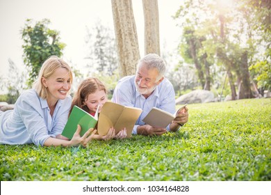 Portrait Of Happy Family Senior Man Woman And Little Girl Reading Outside. Mature Elder Family Of Three Having Fun Reading Book Together Outdoor.  Home School Education Family Lifestyle Spring Summer 