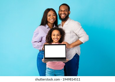 Portrait of happy family of three African American people showing laptop with white empty screen, smiling girl holding gadget with blank space for mock up, standing over blue studio background