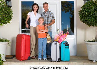 Portrait Of A Happy Family Of Three With Luggage By The Front Door Of House Looking At The Camera.