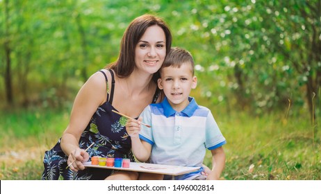 Portrait happy family  Young woman   little boy looking at camera happily  Mom   son drawing pictures together in green park  Horizontal color photography 
