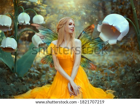 Portrait of happy fantasy woman blonde forest fairy. Fashion model in a bright yellow dress with butterfly wings sits posing in nature. Large flowers scenery decor white lilies. Light magic radiance