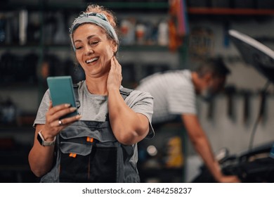 Portrait of happy female auto mechanic workshop worker standing at mechanic workshop with cellphone in hands and smiling at it while having video call. In blurry background is repairman fixing car. - Powered by Shutterstock