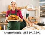 Portrait of happy female baker working in bakehouse, holding tray with fresh bakery goods on light kitchen