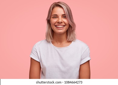 Portrait of happy female with broad smile, wears brackets on teeth and casual white t shirt, poses against pink background. Attractive woman with bobbed hairstyle, being in good mood after date