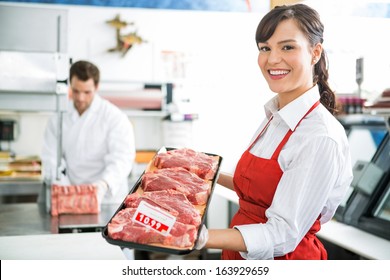 Portrait of happy female butcher holding meat tray in store with colleague working in background - Powered by Shutterstock