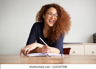 Portrait Of Happy Female College Student Sitting At Desk Writing In Book