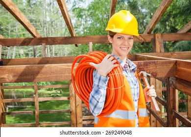 Portrait Of Happy Female Construction Worker Holding Pipe And Hammer In Timber Cabin At Site