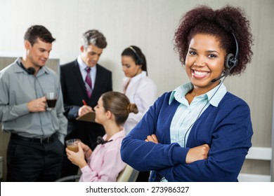 Portrait Of Happy Female Customer Service Representative Standing Arms Crossed While Team Discussing In Background At Office