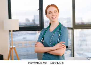 Portrait Of Happy Female Doctor In Blue Green Uniform Standing Arms Crossed Near Window In Sunny Day In Medical Clinic Office. Redhead Woman Surgeon Posing With Stethoscope Looking Away.