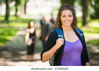 Portrait Of Happy Female Grad Student With Backpack Standing At Campus With Friends In Background