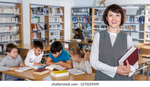 Portrait Of Happy Female Librarian Standing With Book In Hands In School Library 