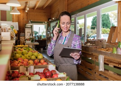 Portrait of happy female sales assistant in a grocery using digital tablet and talking on cell phone while taking inventory - Owner with digital tablet preparing online grocery order - Powered by Shutterstock