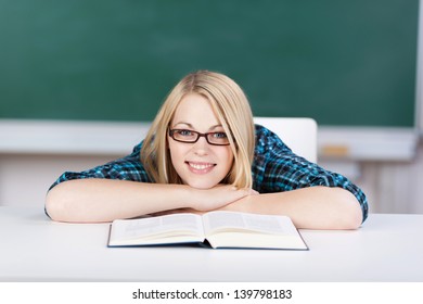 Portrait of happy female student with book leaning on desk in classroom