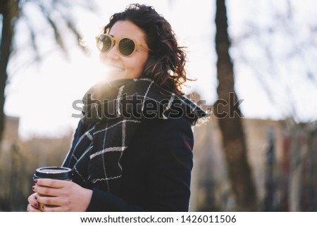 Portrait of happy female student enjoying coffee break on urban setting laughing during sunny autumn day, positive cheerful hipster girl in trendy sunglasses and scarf holding paper takeaway cup