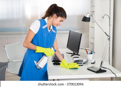 Portrait of happy female worker cleaning computer desk with spray and sponge at office