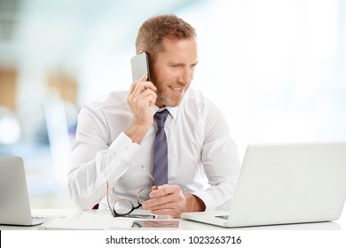 Portrait of happy financial assistant businessman using mobile phone and making call while working on laptop at the office.  - Powered by Shutterstock