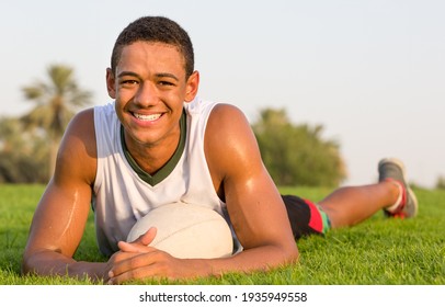 Portrait of happy fit African teen athlete relaxing on the grass with a rugby ball and smiling to camera.  - Powered by Shutterstock