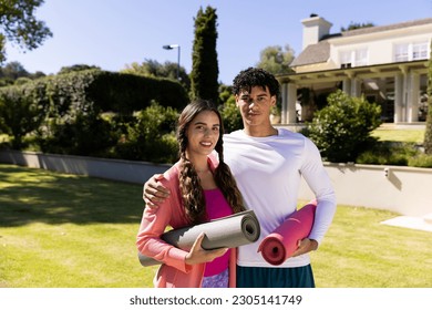 Portrait of happy fit diverse couple standing in sunny garden embracing and holding yoga mats. Summer, healthy lifestyle, relaxation and vacations. - Powered by Shutterstock