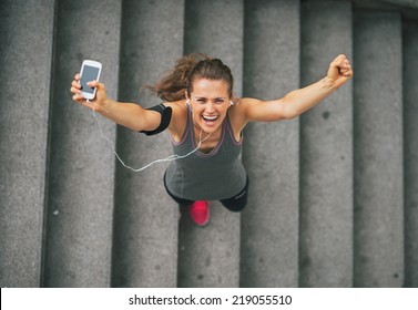 Portrait of happy fitness young woman with cell phone outdoors in the city rejoicing