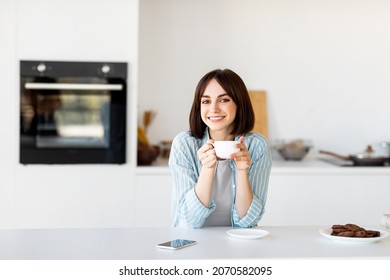 Portrait of happy freelancer lady enjoying cup of hot coffee in kitchen and smiling at camera, free space. Excited millennial woman sitting at table, having break in work, copy space