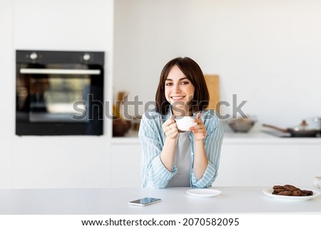 Portrait of happy freelancer lady enjoying cup of hot coffee in kitchen and smiling at camera, free space. Excited millennial woman sitting at table, having break in work, copy space
