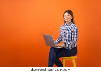 Portrait of happy freelancer, young woman with brown hair in checkered casual shirt and denim sitting, working on laptop and smiling at camera. indoor studio shot isolated on orange background - Shutterstock ID 1575490774