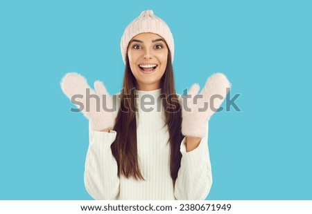Portrait of happy friendly laughing girl in pink hat, mittens and white sweater waving hands gesturing hi, hello on blue background looking at camera. Winter, warm style. Sincere emotions concept.