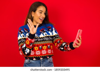 Portrait Of Happy Friendly Young Beautiful Hispanic Woman Wearing Winter Sweater Against Red Wall  Taking Selfie And Waving Hand, Communicating On Video Call, Online Chatting.