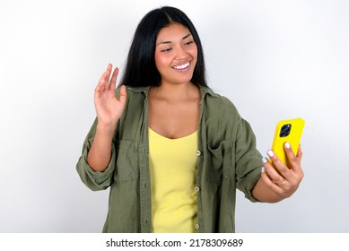 Portrait Of Happy Friendly Young Hispanic Woman Wearing Green Jacket Over White Background Taking Selfie And Waving Hand, Communicating On Video Call, Online Chatting.