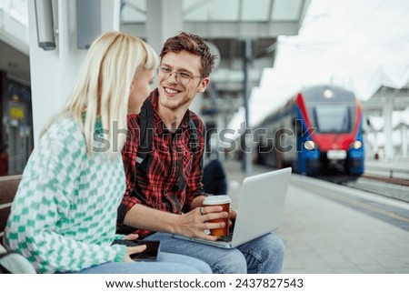 Portrait of a happy friends or a couple sitting on a bench at the railway station and drinking coffee and using a laptop. Man and woman waiting for a train to arrive and spending their time together.