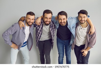 Portrait Of Happy Friends Standing With Arms Around Each Other's Shoulders In Studio With Light Gray Background. Group Of Young Bearded Men Hugging, Smiling, Looking At Camera And Having Fun Together