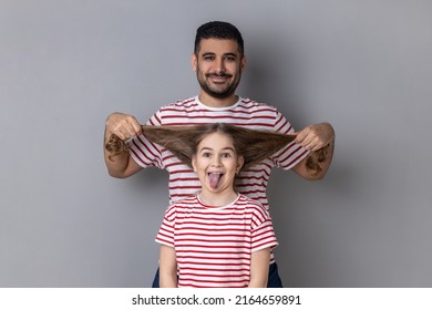 Portrait Of Happy Funny Father And Daughter Having Fun, Cute Kid Showing Tongue Out, Dad Pulling Her Hair, Expressing Positive Emotions. Indoor Studio Shot Isolated On Gray Background.