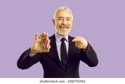 Portrait Of Happy Funny Old Man In Suit Isolated On Violet Studio Background Hold Point At Casino Chips Recommend Easy Win. Smiling Mature Male Poker Player Or Croupier Encourage Gambling.