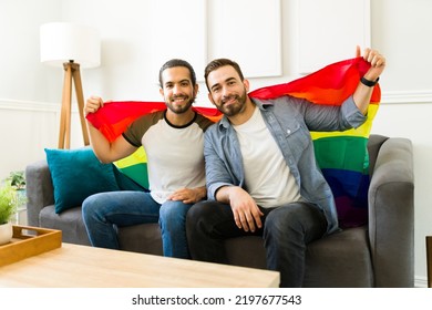 Portrait Of A Happy Gay Man And His Partner Supporting LGBT Rights And Waving A Rainbow Flag While At Home