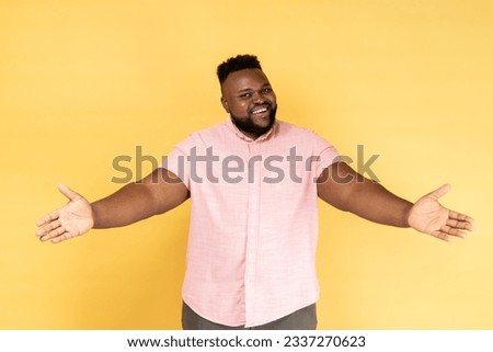 Portrait of happy generous bearded man wearing pink shirt standing with raised hands and looking at camera, welcoming or giving smth. Indoor studio shot isolated on yellow background.