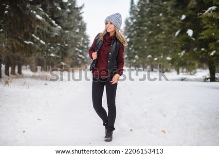 portrait of a happy girl in a blue hat, sweater and vest, stands on a snowy winter alley among the Christmas trees