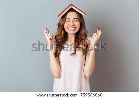 Portrait of a happy girl holding book on her head with crossed fingers for good luck isolated over gray wall background