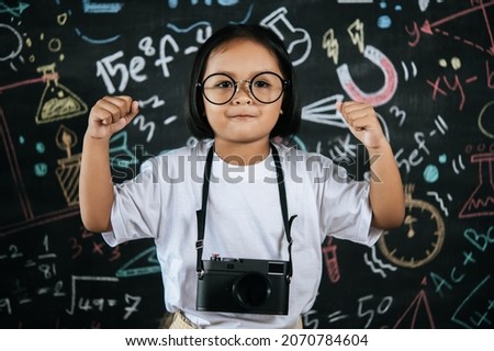 Portrait of happy girl holding digital camera, Little photographer girl enjoy to use camera take a photo, she standing at front of blur blackboard in background