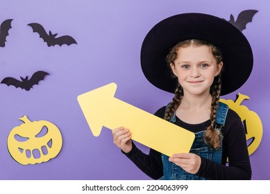 Portrait Of Happy Girl Kid In Big Hat Showing Aside With Yellow Arrow, Looking At Camera, Isolated On Purple Background Decorated Paper Pumpkins And Bats With Copy Space For Advertisement. Halloween