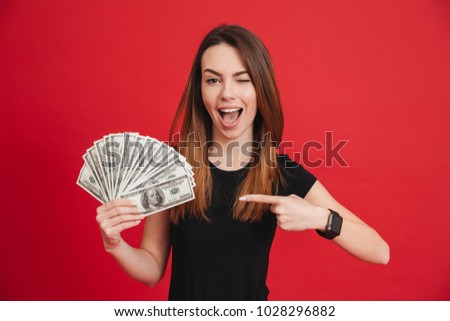 Portrait of a happy girl pointing finger at bunch of money banknotes and winking isolated over pink background