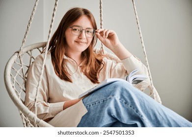 Portrait of happy girl relaxing and reading book on indoor swing. Pretty female in glasses excited about story she reading. Cozy afternoon with book in hanging chair in home. - Powered by Shutterstock